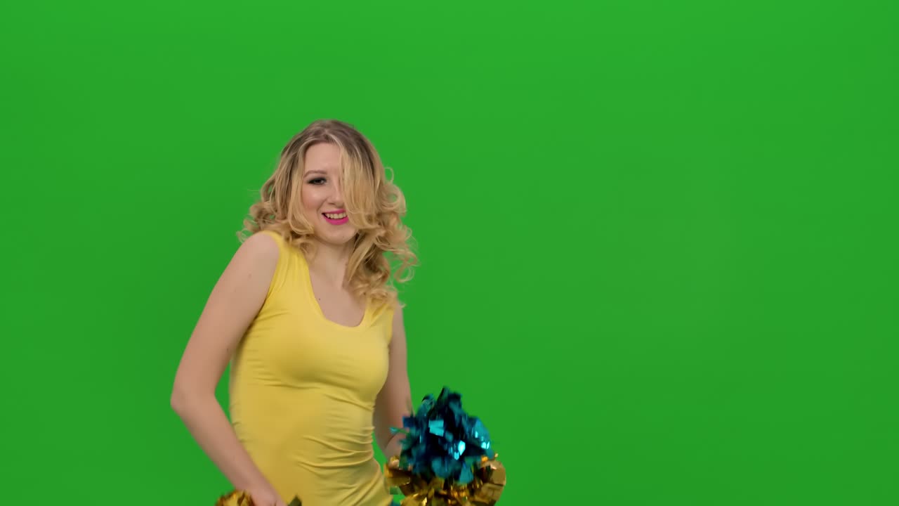 치어리더는 허지, 폼폼 팔, 머리카락을 돌리고 흔들며 활기차게 움직이고, 노란색과 파란색의 유니폼을 입은 소녀가 녹색 스크린에서 스튜디오에서 즐거운 춤을 추고 있습니다.