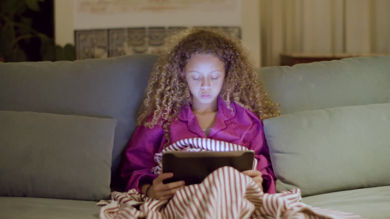 niña en pijama sentada en el sofá leyendo un libro electrónico