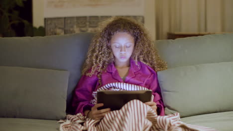 Young-girl-in-pyjamas-sitting-on-couch-reading-ebook