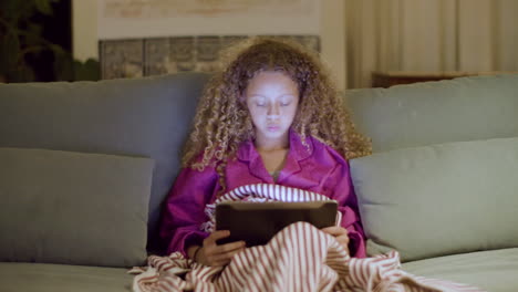 niña en pijama sentada en el sofá leyendo un libro electrónico