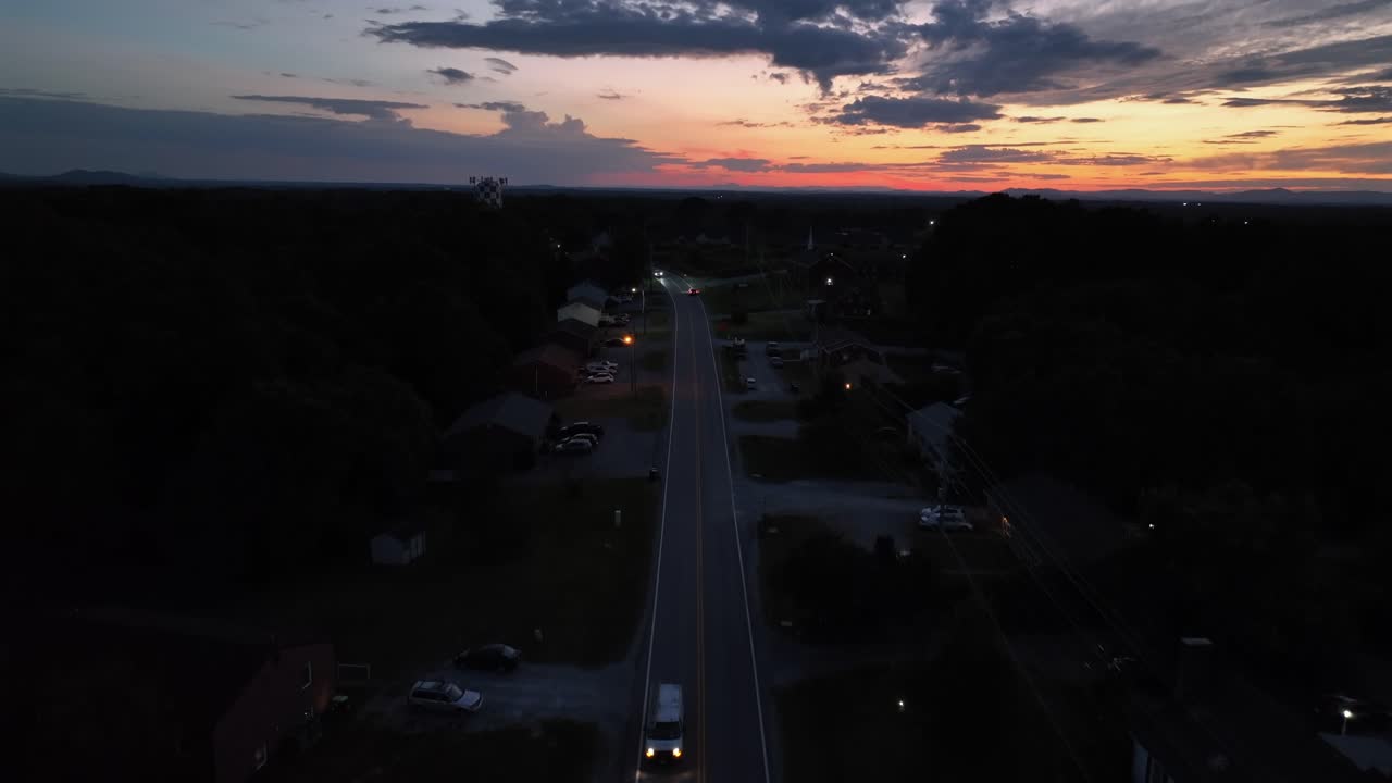 Driving vehicle on suburb road of dark neighborhood during golden sunset. Peaceful night scene. Aerial rising wide shot. Houses and homes in quaint city of USA