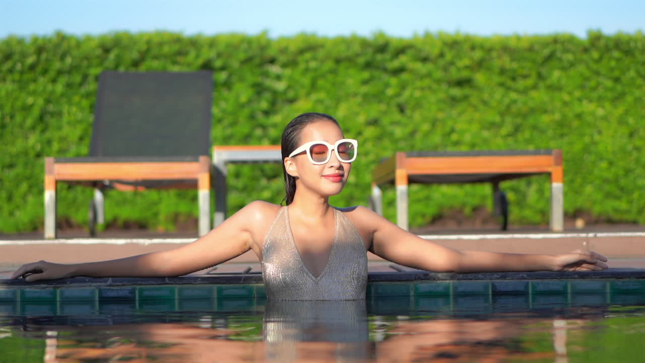 반짝이는 금색 수영복과 수영장에 서 있는 큰 선글라스를 쓴 매력적인 여성이 고개를 돌려 매혹적인 미소로 카메라를 바라보고 있습니다.
