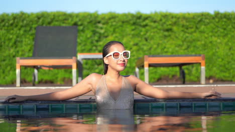 mujer atractiva en traje de baño dorado brillante y grandes gafas de sol de pie en la piscina gira la cabeza y mira hacia la cámara con una sonrisa seductora