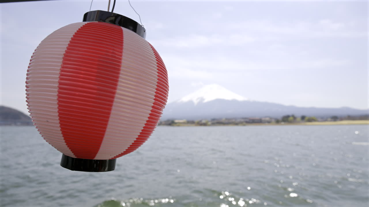 A vibrant lantern with red and white colors sways gently, offering a beautiful contrast against the stunning backdrop of Mount Fuji reflected over the calm lake Kawaguchi