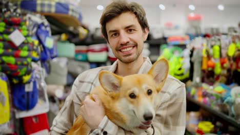 Portrait-of-a-happy-brunette-guy-in-a-plaid-shirt-who-holds-his-yellow-corgi-dog-in-his-arms-in-a-pet-store