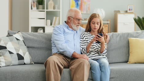 Grandfather-Embracing-His-Pretty-Granddaughter-While-They-Sitting-On-The-Sofa-In-The-Living-Room-And-Girl-Tapping-On-The-Smartphone-Screen-And-Man-Watching-At-It