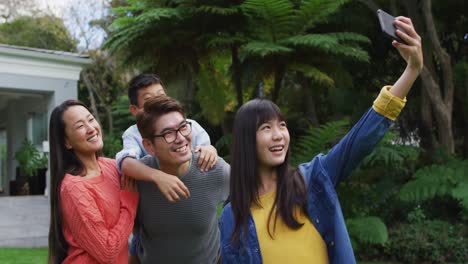 Happy-asian-daughter-taking-selfie-with-happy-mother,-and-father-piggybacking-brother,-in-garden