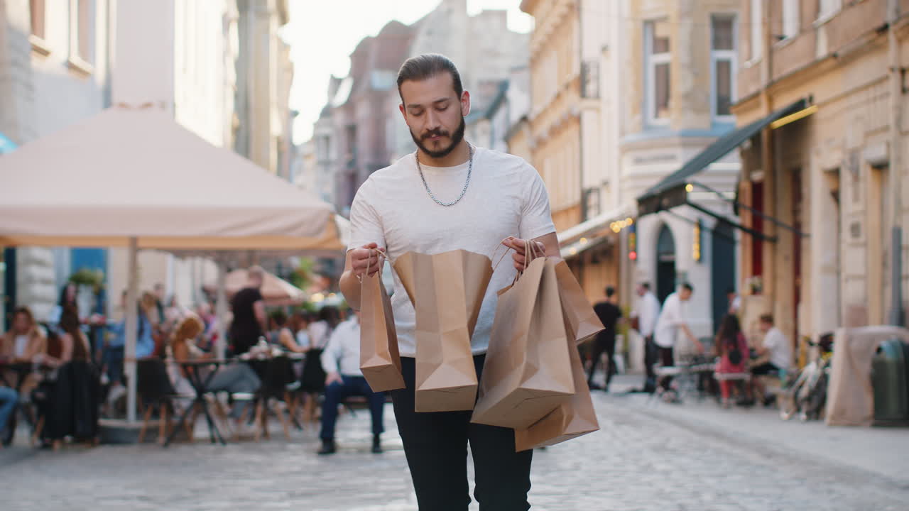 Un giovane uomo felice, un consumatore appassionato di shopping, dopo la vendita degli acquisti con le borse piene, cammina per la strada della città.