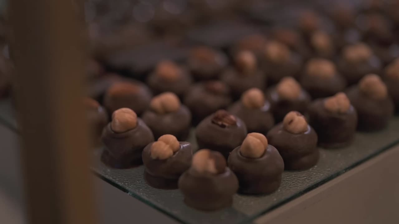 Closeup shot scanning a large tray of assorted artisan chocolates, featuring round pieces with cashews and almonds, and long bars with red decorative lines