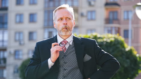 Portrait of mature businessman holding eyeglasses outdoors. Middle aged man with grey hair wearing elegant suit and glasses standing in the street and telling something happily.
