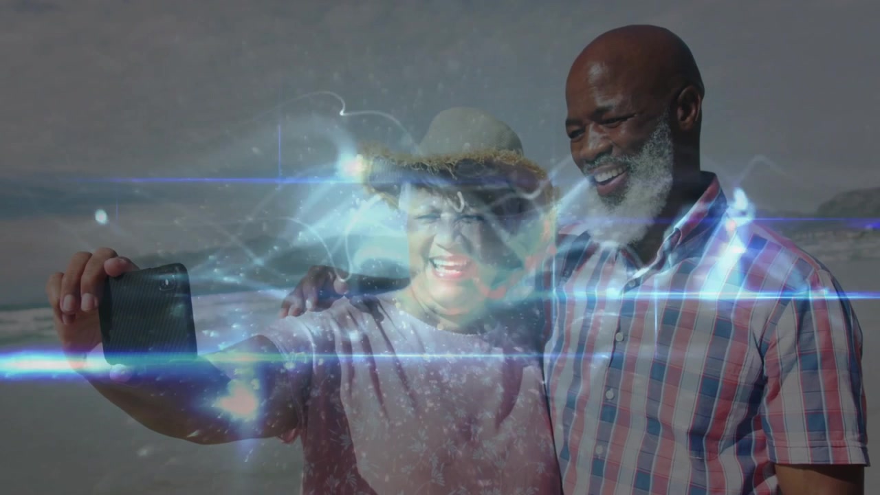 Senior couple raising phone for selfie while light streaks showing technology on beach