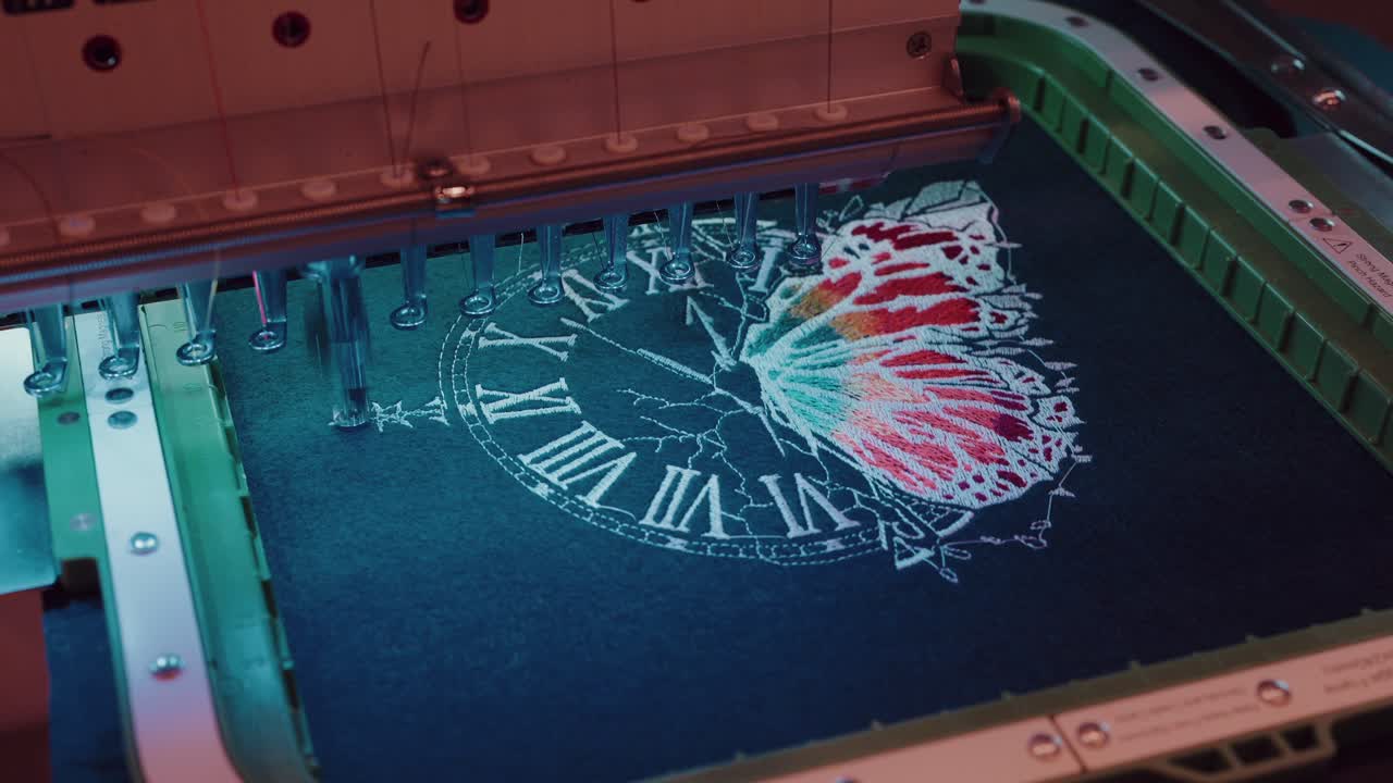 Embroidery machine stitching a colorful butterfly and clock design on dark textile fabric