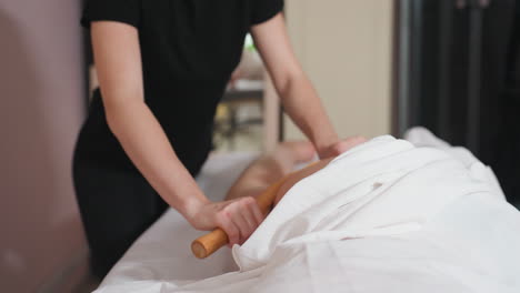 massage therapist in black using wooden massage stick to gently massage client thigh on white-covered table inside softly lit treatment room, focusing on muscle relaxation and therapeutic care