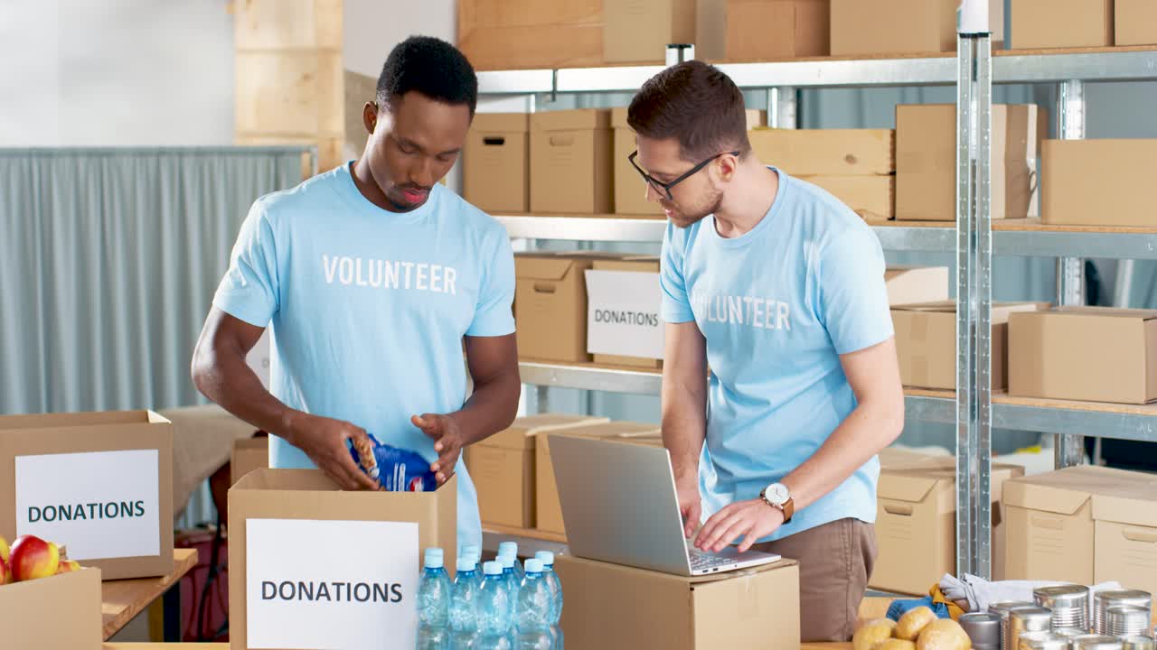 voluntario afroamericano empacando una caja de donación con comida en un almacén de caridad mientras su compañero de trabajo escribe en una laptop