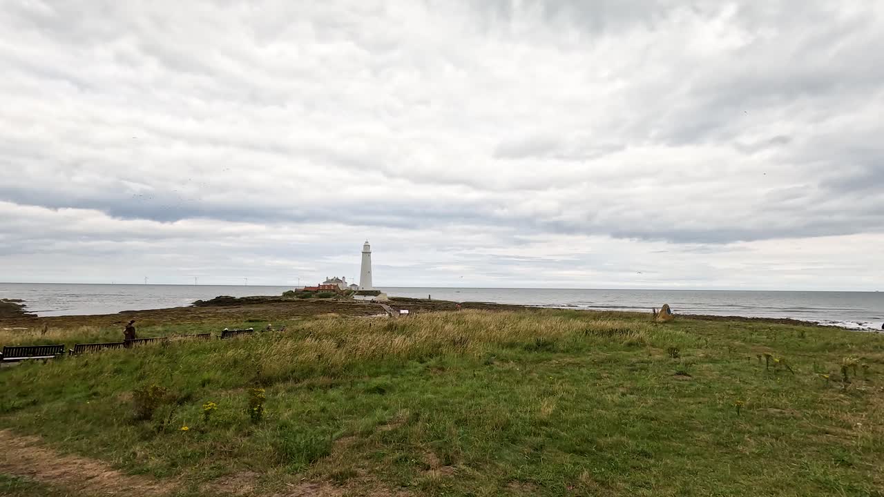 A wide-angle camera pan reveals a windswept grassy coastline with benches, leading to a distant white lighthouse under overcast skies and soft daylight