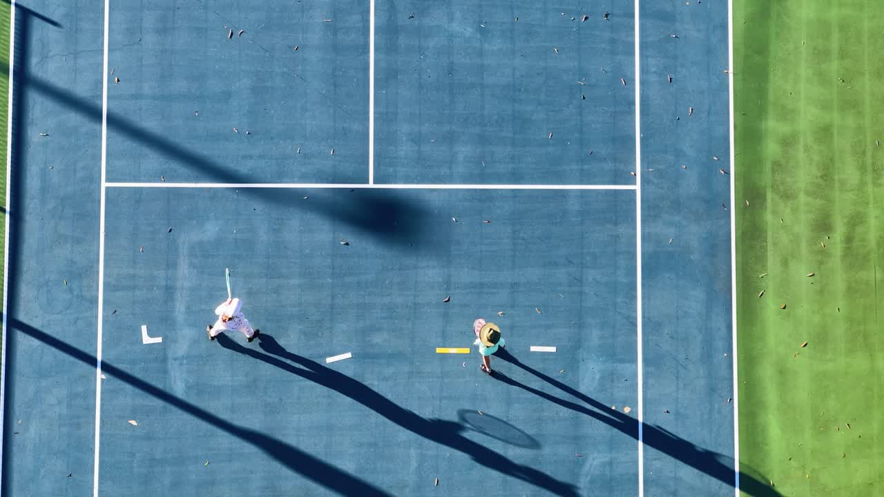 Two players engage in a tennis match on a sunny day, captured from above with dynamic shadows