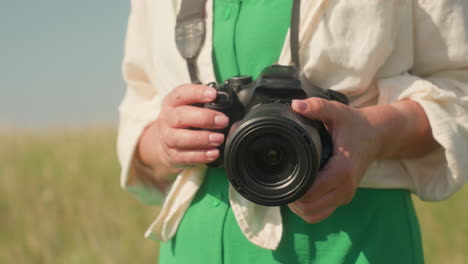 close up of woman in green dress and cream shirt walking with camera in hands, natural daylight illuminating camera lens and fingers, blurry countryside landscape in background