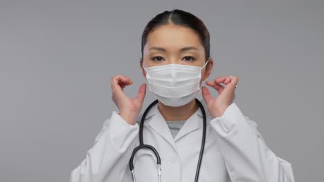 Glückliche-Asiatische-Ärztin-Zeigt-Medizinische-Maske.Medizin-,-Berufs--Und-Gesundheitskonzept-–-Videoporträt-Einer-Glücklich-Lächelnden-Asiatischen-Ärztin-Im-Weißen-Kittel-Mit-Stethoskop,-Die-Zeigt,-Wie-Man-Eine-Medizinische-Maske-Auf-Grauem-Hintergrund-Verwendet