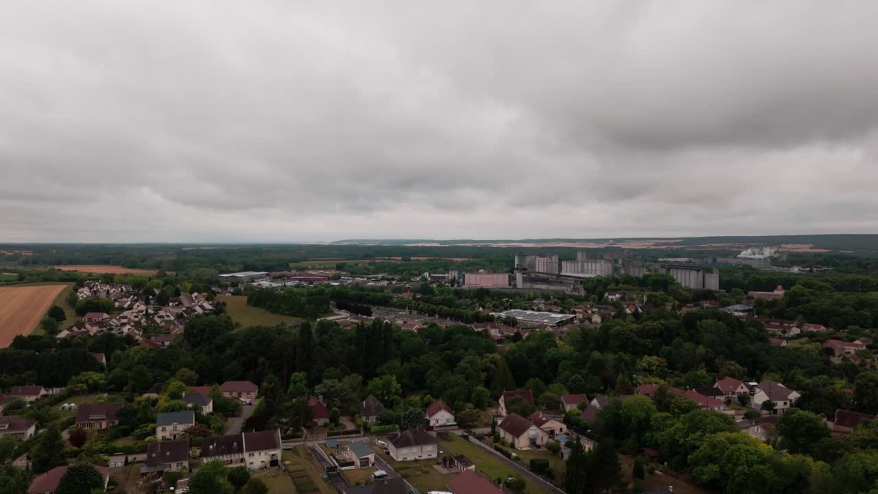 High angle drone view over a suburban French town, showing a mix of residential houses, apartment buildings, green trees, and distant fields under an overcast sky