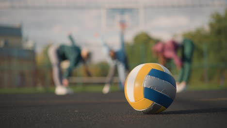 close-up of volleyball on outdoor court with blurred view of athletes bending during workout in background, building and sports equipment visible, vibrant sunlight illuminating scene