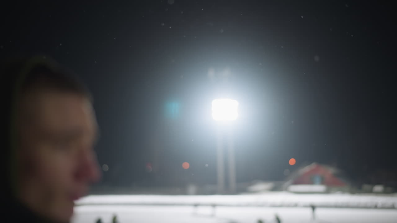 Bright overhead stadium light illuminates dark winter night as snowflakes drift gently through sky, creating dramatic moody atmosphere with particles floating in foreground against black background