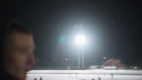 bright overhead stadium light illuminates dark winter night as snowflakes drift gently through sky, creating dramatic moody atmosphere with particles floating in foreground against black background