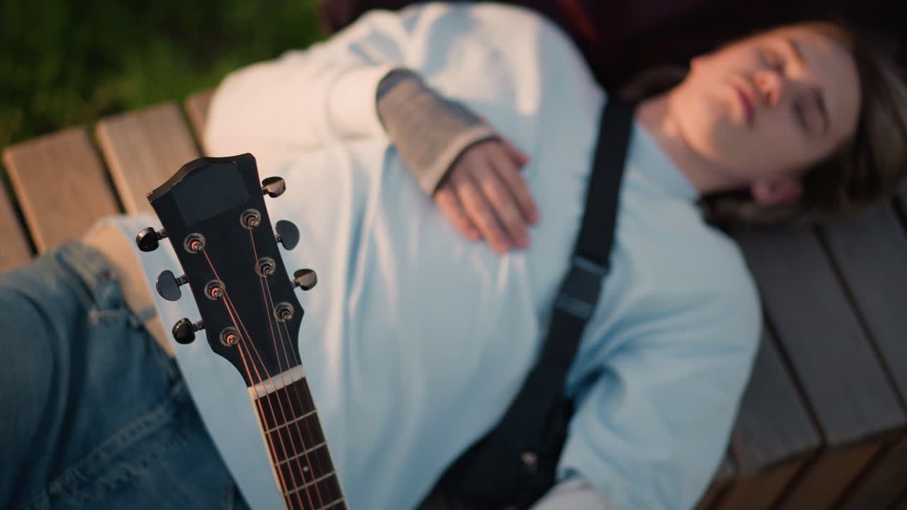 white guitarist lying on wooden bench beside acoustic guitar, wearing white sweater and fingerless gloves, strap across chest, eyes closed in golden light, relaxed songwriting mood in park