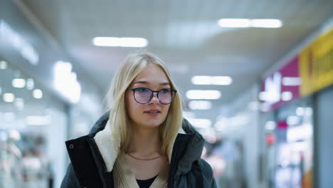 young lady in glasses and black hoodie walking through well-lit mall, looking around with a soft smile, blurred background with other shoppers and glowing lights creating a bokeh effect