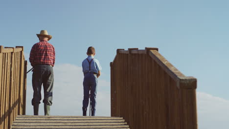 rear view of senior man in a hat standing on the wooden bridge with little boy and fishing with rods in hands