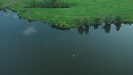 expansive green lakeside bank lined with trees and grassy shore reflecting crescent moon and drifting cloud shapes under soft dusky sky creating tranquil rural evening