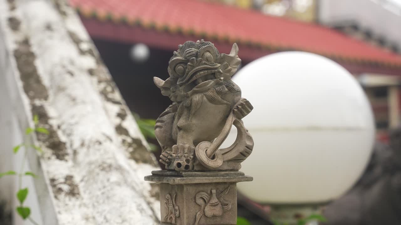 A tight zoom centers on a fierce stone creature with curled mane and paw on a ball. The red-roofed temple and white sphere blur behind, spotlighting texture, strength, and symbolic presence