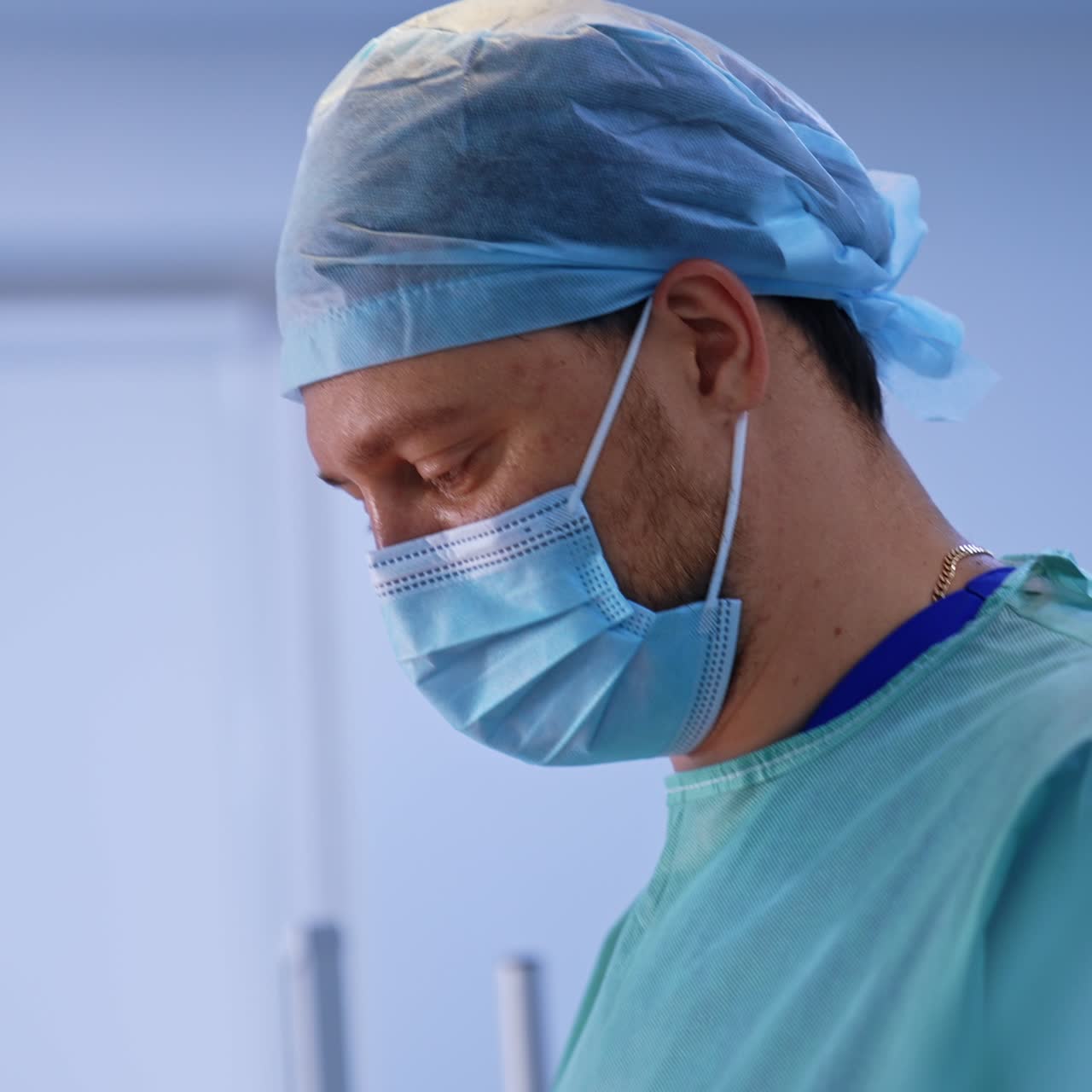 Doctor in cap and mask working in modern surgery room. Side portrait of a male surgeon focused on his work. Blurred backdrop