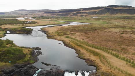 the less crowded but beautiful waterfall glanni seen from above, surrounded by a magical icelandic landscape with lush green bushes and majestic mountains in the background