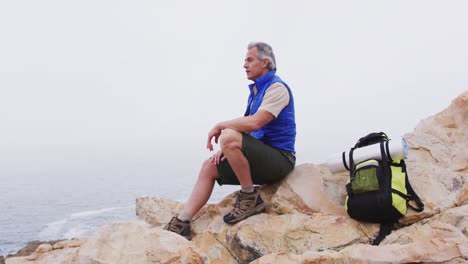 Hombre-Excursionista-Mayor-Con-Mochila-Sentado-En-Las-Rocas-De-Las-Montañas-Y-Mirando-La-Orilla-Del-Mar.