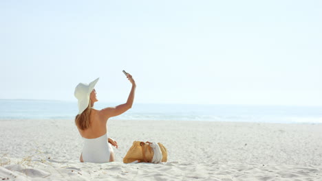 Frau,-Die-Mit-Dem-Telefon-Ein-Selfie-Macht-Und-Am-Strand-Sitzt-Und-Einen-Einteiligen-Designer-Badeanzug-Trägt