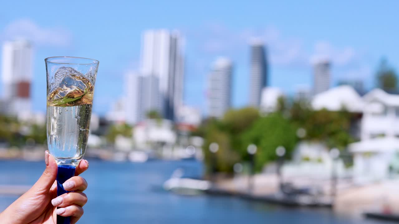 A hand gently swirls a champagne glass, creating motion in the liquid, with a sunny waterfront cityscape and high-rise buildings softly blurred in the background