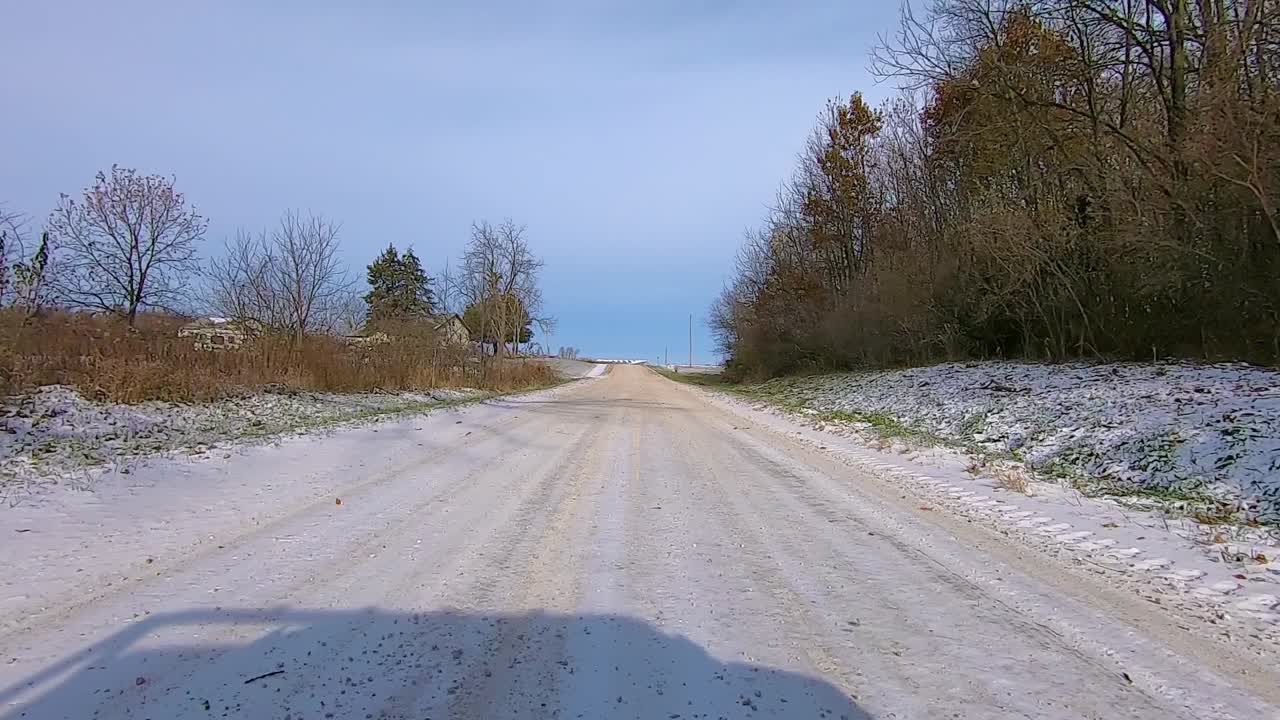 POV through the rear window while driving on a rural gravel road past farm houses and timber on an early winter afternoon; backwards Point of View