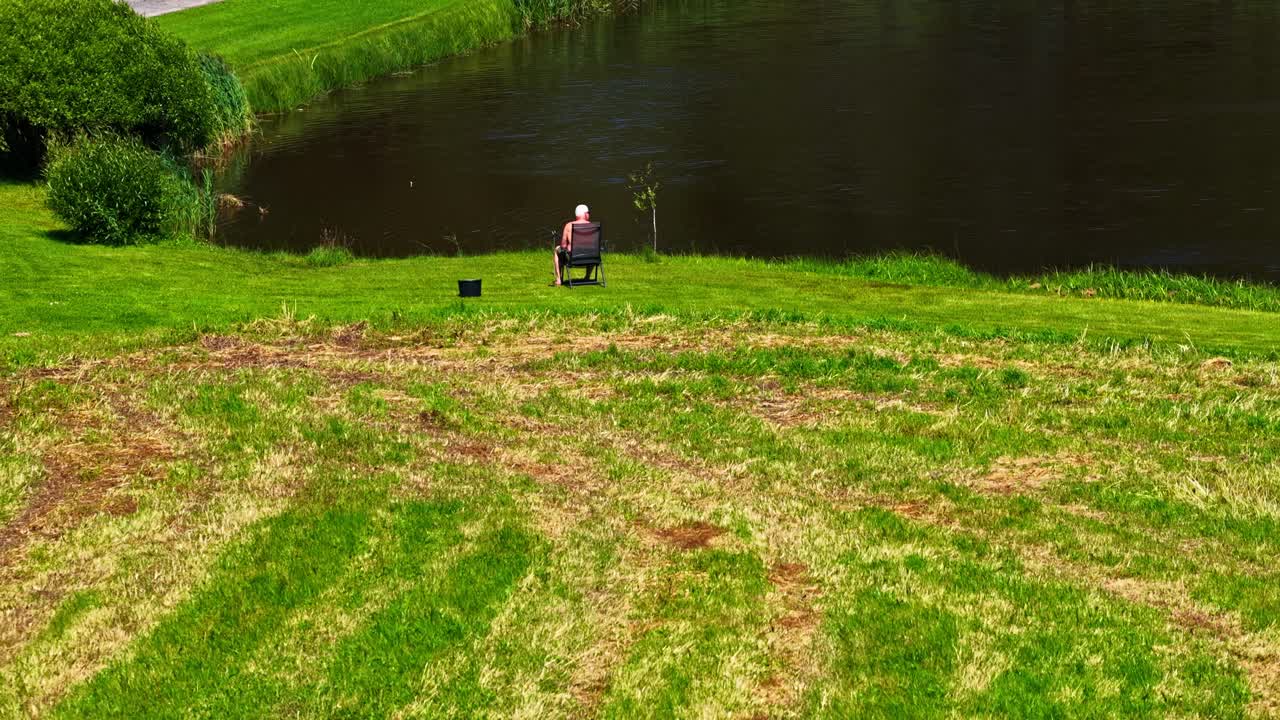 Aerial pullback of elderly man sitting alone beside a green pond on rural lawn