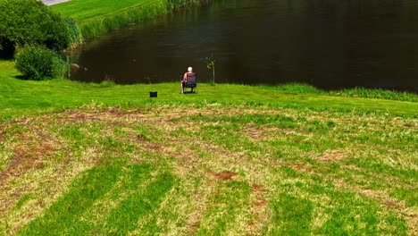 aerial pullback of elderly man sitting alone beside a green pond on rural lawn