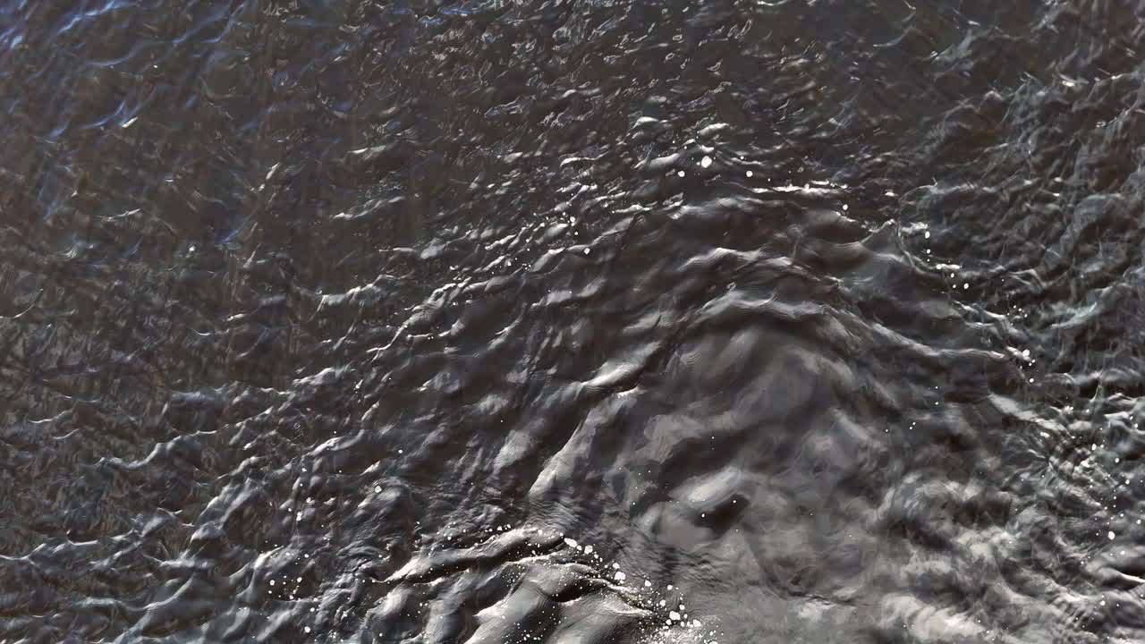 An arc of white bubbles drifts across rippling brown floodwaters, suggesting underwater turbulence and natural force. The textured surface creates an abstract, dramatic visual