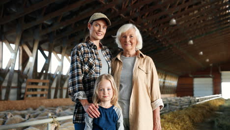 Portrait-of-Caucasian-grandmother,-mother-and-little-girl-smiling-at-camera-in-a-stable-with-sheep-flock