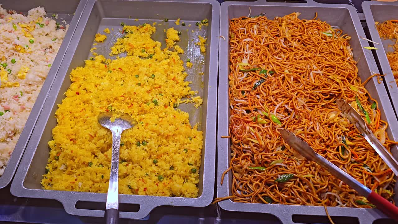 Camera pans sideways across buffet trays with noodles, rice, and broccoli
