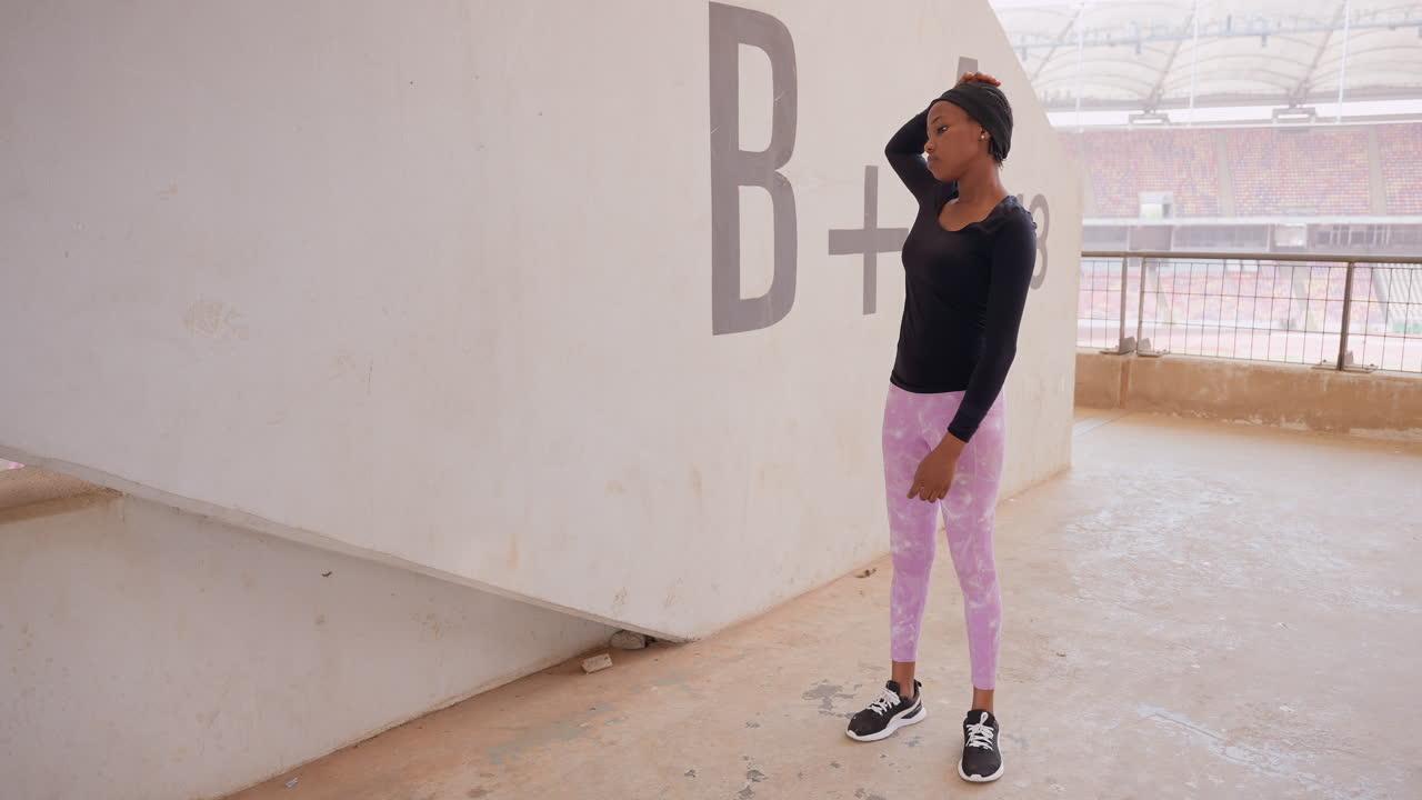 Mujer negra posando cerca de una pared de hormigón. Modelo vistiendo top negro y pantalones rosas con zapatillas, alternando entre poses pensativas y seguras, con una mano apoyada en la cabeza, apoyada contra una superficie texturizada.