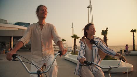 Front-view-of-a-happy-couple,-a-guy-and-a-girl,-ride-bicycles-along-the-beach-on-which-palm-trees-grow-near-the-sea-at-sunrise-in-summer.-Beautiful-modern-beach,-guy-and-girl-riding-bicycles