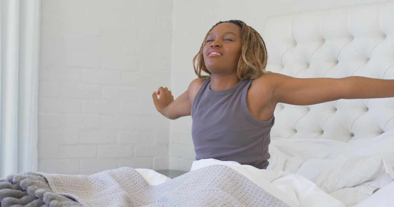Donna afroamericana che si sveglia e si stira a letto con una testiera imbottita, abbassando le braccia e sistemando la coperta.