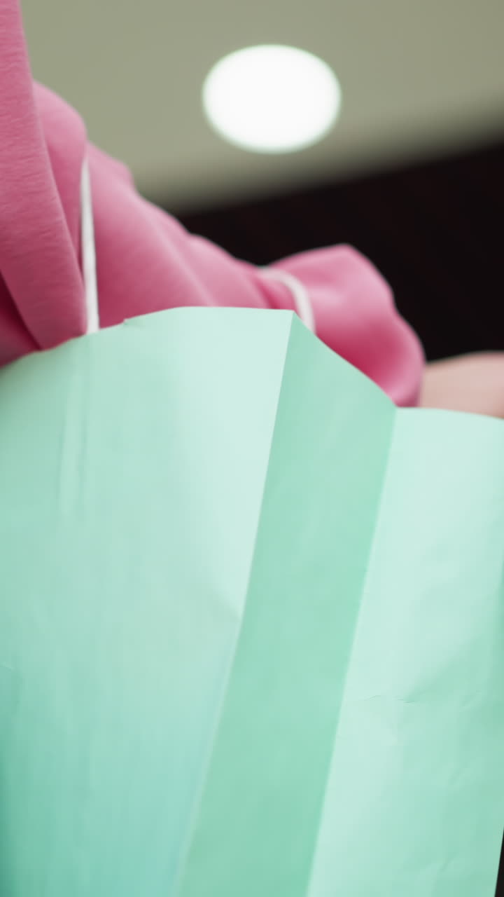 vista de la mano de la dama blanca en vestido rosa sosteniendo una bolsa de compras verde menta con una bolsa negra, ambas manos descansando en el riel de la escalera mecánica mientras se mueve hacia arriba, las luces borrosas son visibles en el fondo