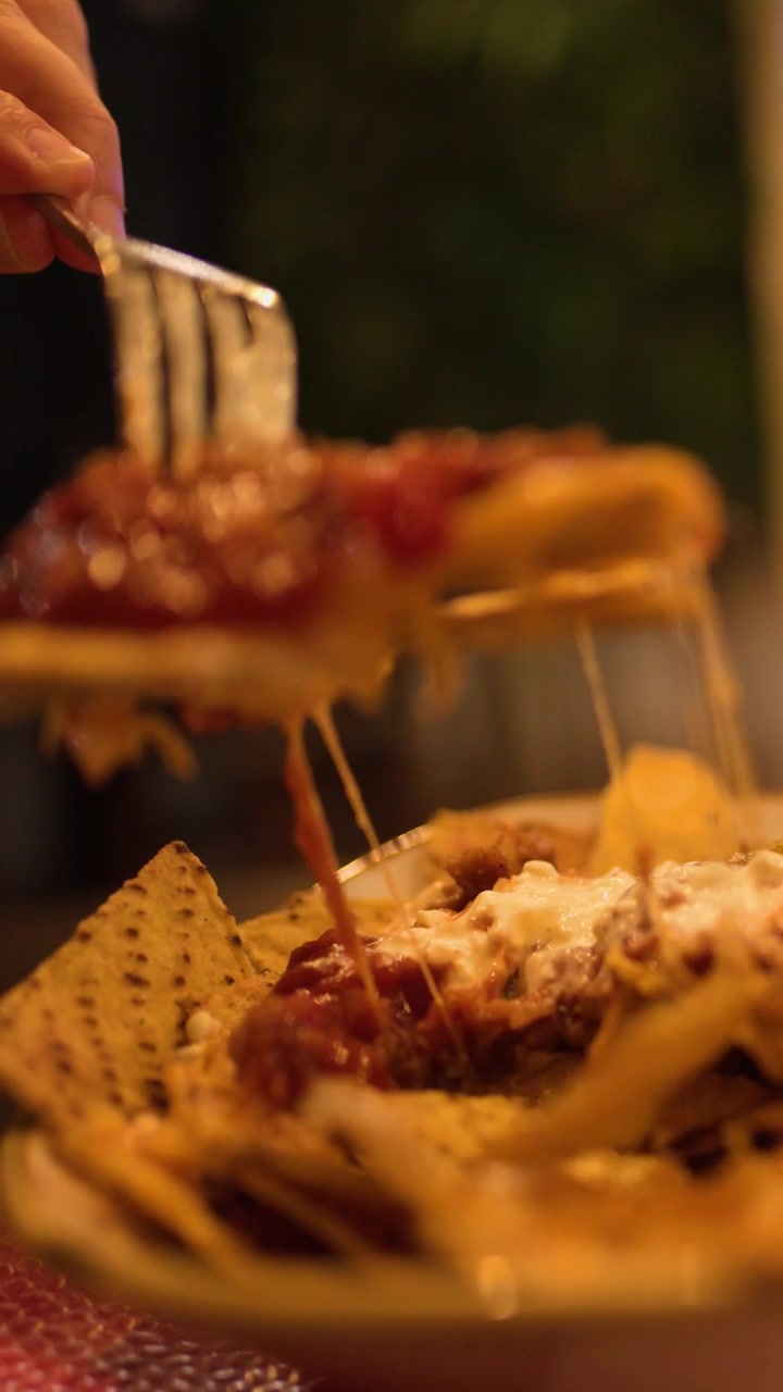 Una mano solleva nachos al formaggio con salsa dal piatto, illuminazione interna calda, messa a fuoco ridotta, primo piano.