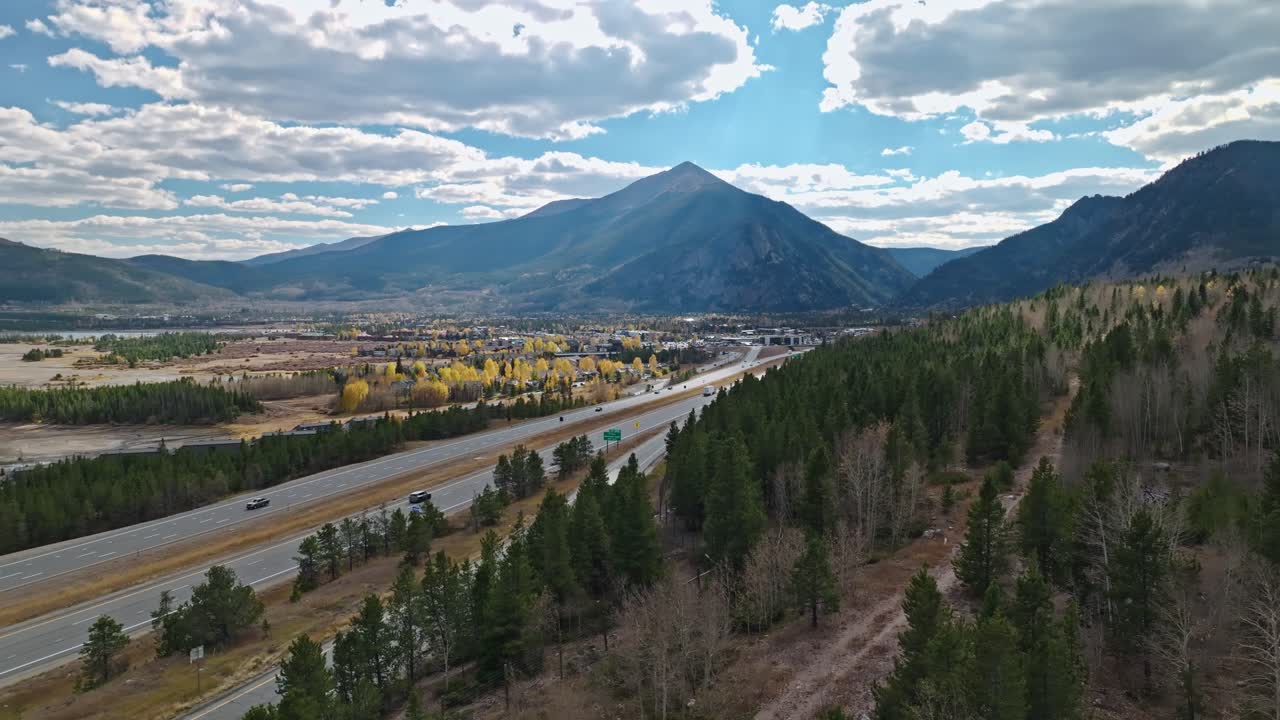 Drone ascends over forest as cars zoom by below distant mountains