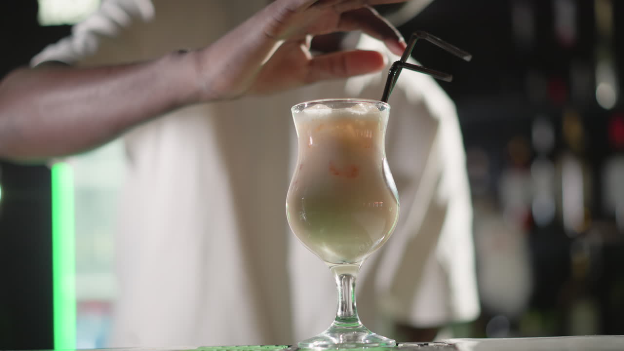 Bartender placing straw into freshly prepared cocktail in glass, adding final touches before serving. Behind, illuminated bar shelves and Heineken neon sign glowing brightly