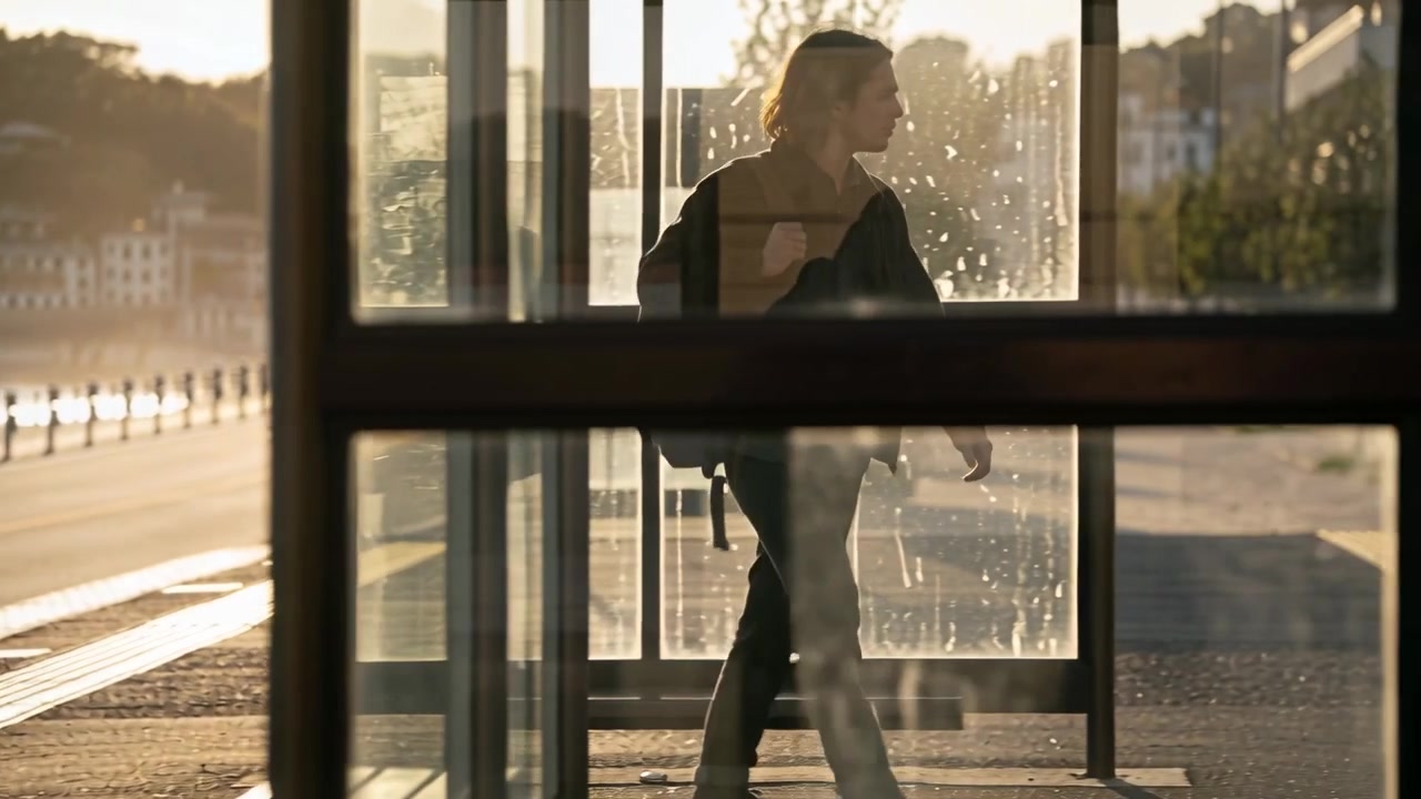 Walking male in dark coat with backpack reaching glass shelter, stepping inside, gazing toward bay